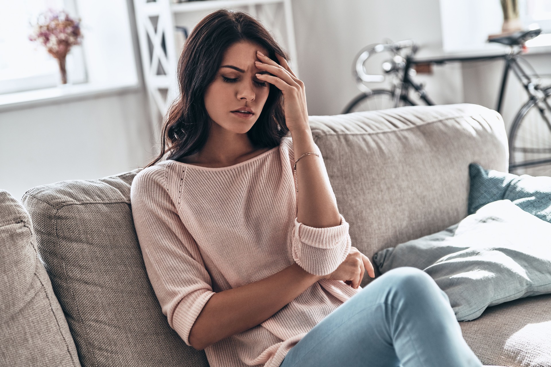 Young woman sitting on a sofa, holding her forehead and looking distressed, possibly experiencing a headache or migraine.